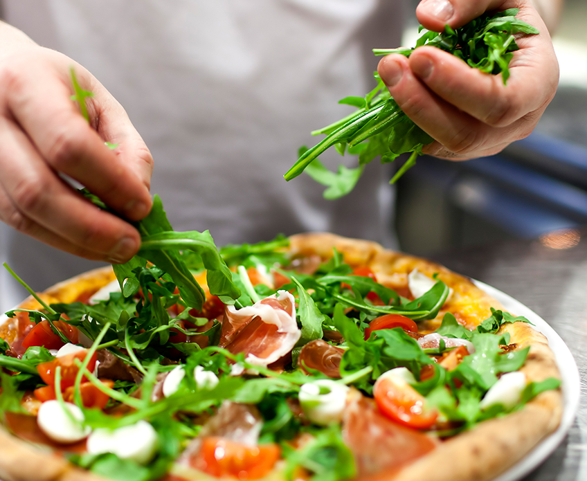 Un chef ajoute de la roquette sur une alléchante pizza Parma garnie de sauce tomate, de mozzarella et de prosciutto.